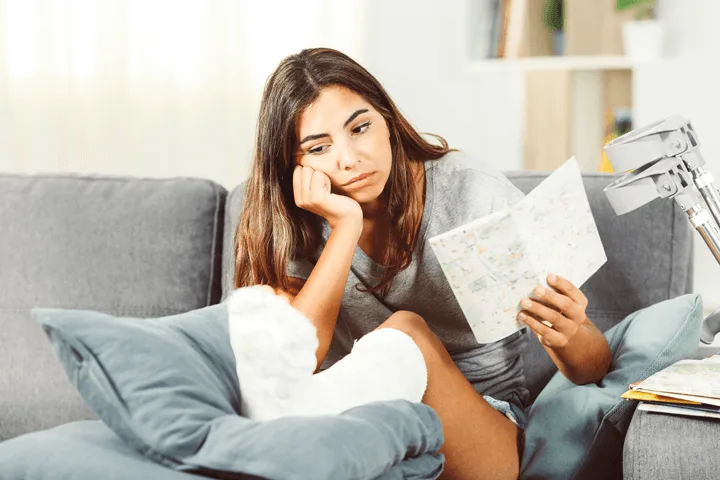 Eine Frau sitzt mit einer Beinverletzung auf dem Sofa. Sie schaut traurig auf eine Landkarte.